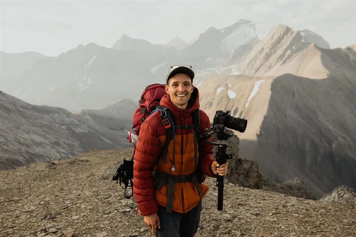 Sam smiling on a mountain ridge, camera rig in hand and hiking pack on his back