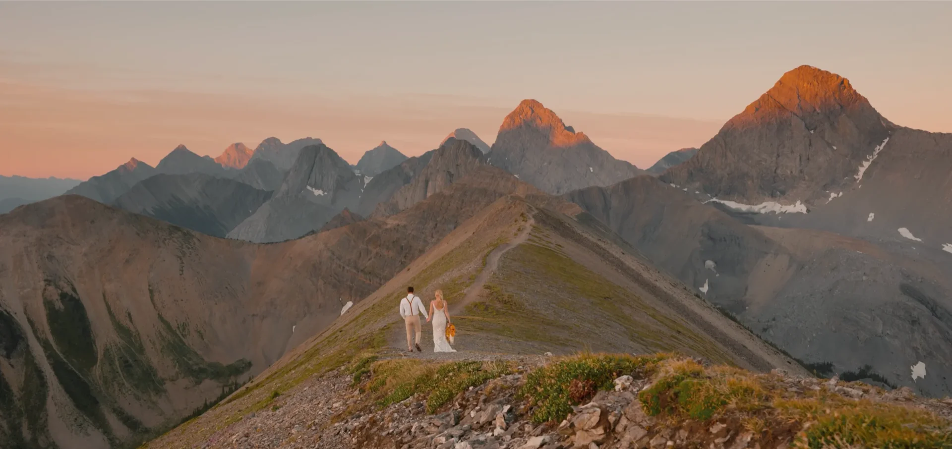 Jayme and Zach walking a grassy ridgeline towards sunlit peaks at dawn