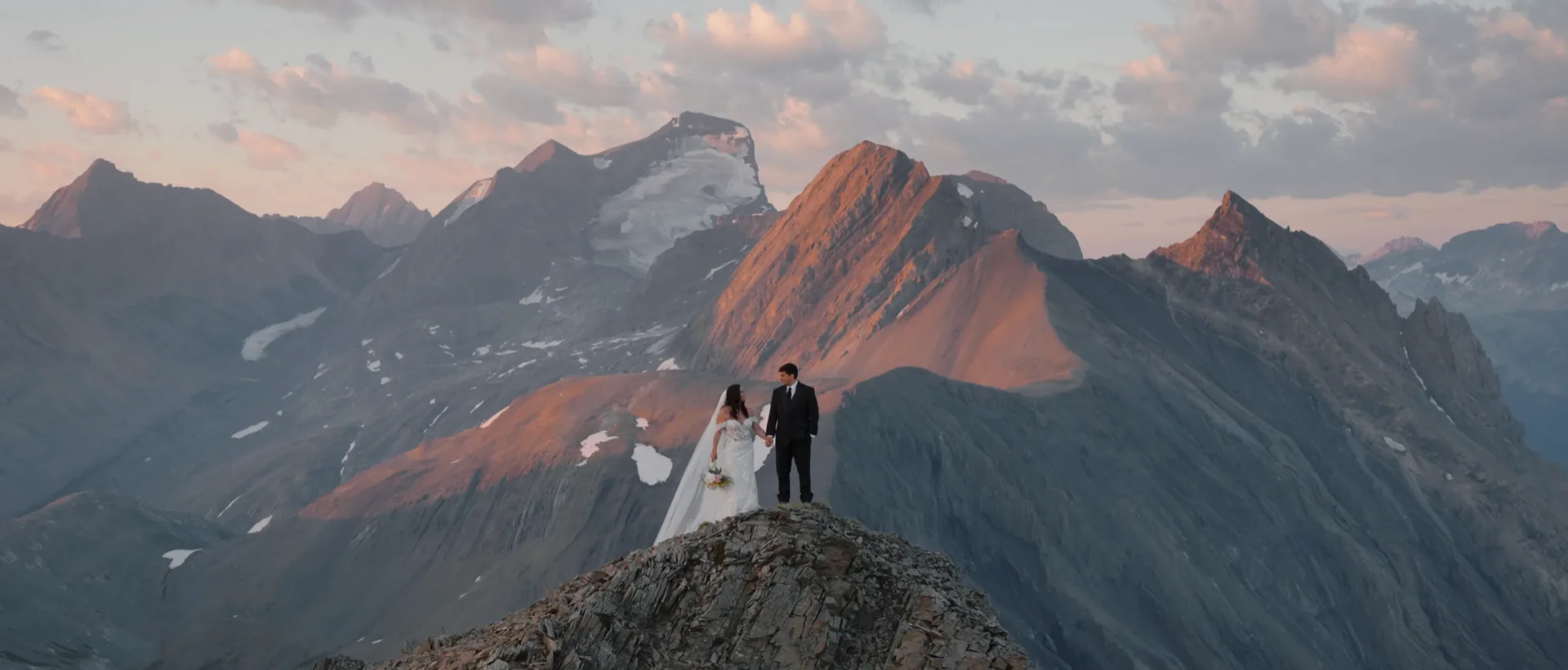 Lexi and Nate standing on a narrow rocky ridge with alpenglow lighting the peaks behind them