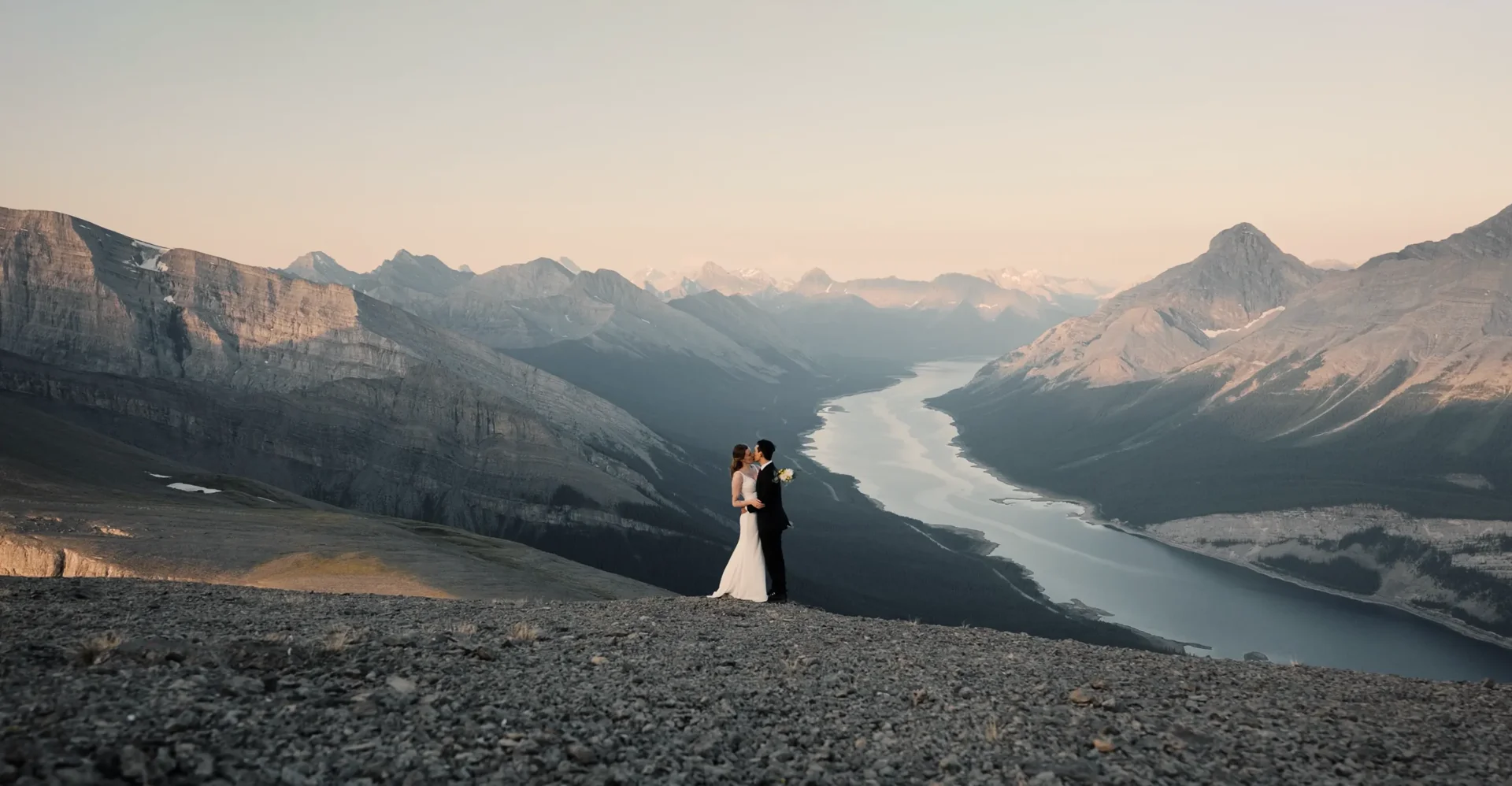 Sarah and Hugo embracing on a mountain summit above a winding turquoise lake at sunrise
